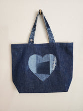 Load image into Gallery viewer, Deep blue denim tote bag with repurposed waistband as handle. The design of bag is m inimalistic wit h a decorative heart sewn in four shades blue of patchwork style.
