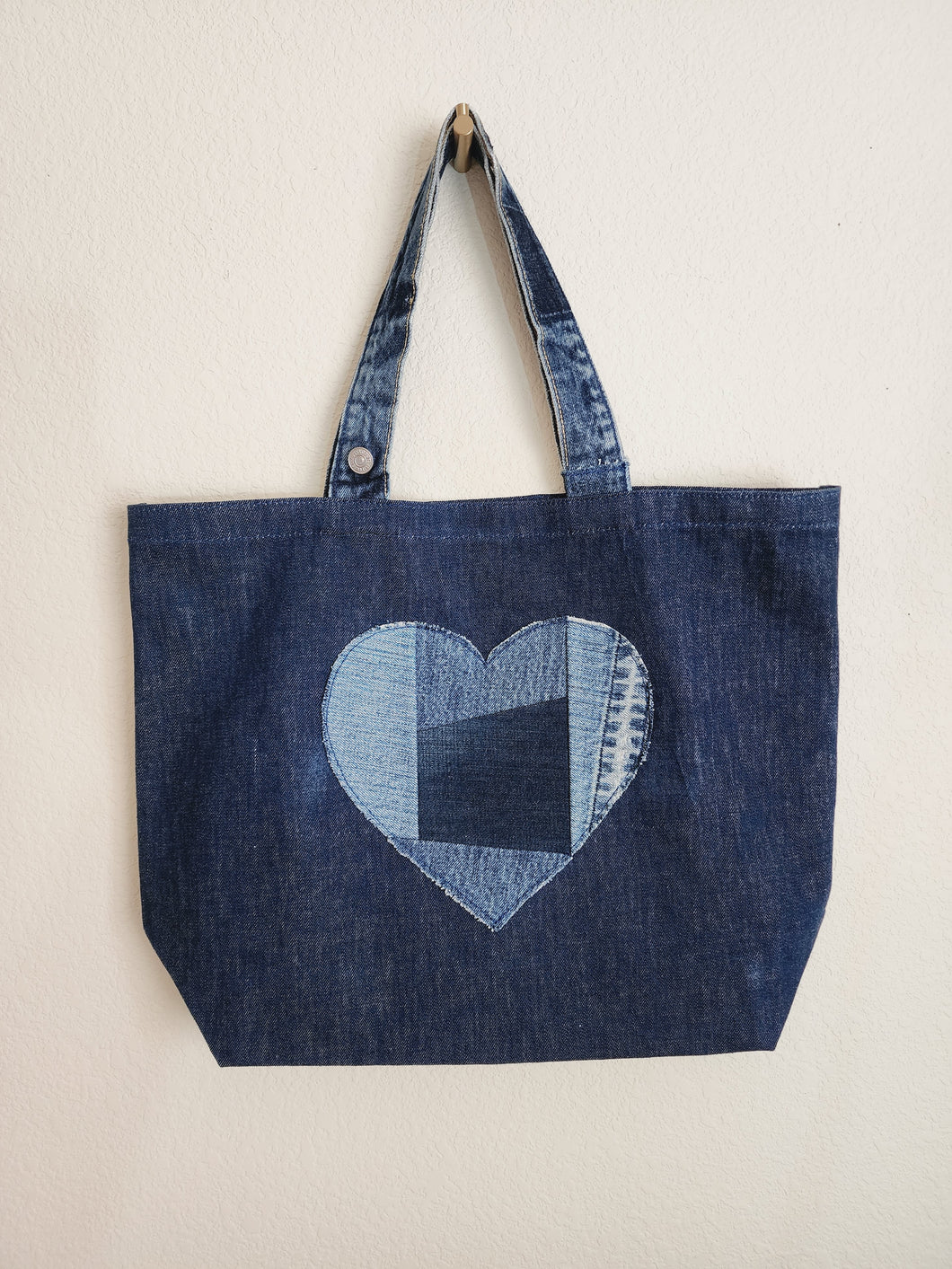 Deep blue denim tote bag with repurposed waistband as handle. The design of bag is m inimalistic wit h a decorative heart sewn in four shades blue of patchwork style.